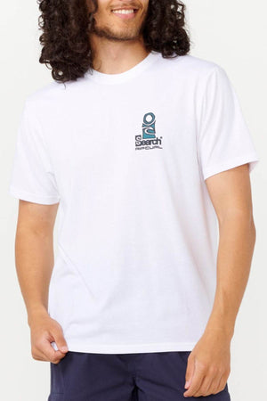 A person with long curly hair is wearing a white Rip Curl Vaporcool Search Stack Tee, displaying a small "Search Rip Curl" logo on the chest. With its moisture-wicking design, it complements dark shorts perfectly. Holding the edge of the shirt in their hand highlights its casual yet functional style.