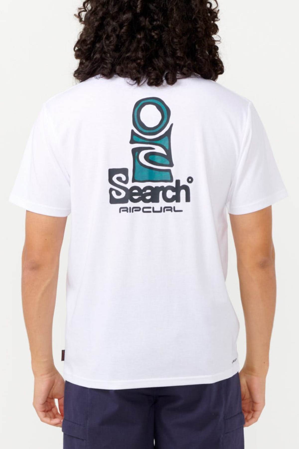A person wearing a white Rip Curl Vaporcool Search Stack Tee, which displays a large "Search" logo on the back with abstract design elements in teal and black. The shirt is designed with quick-drying fabric for all-day comfort. They have long, curly hair and are also wearing dark shorts.