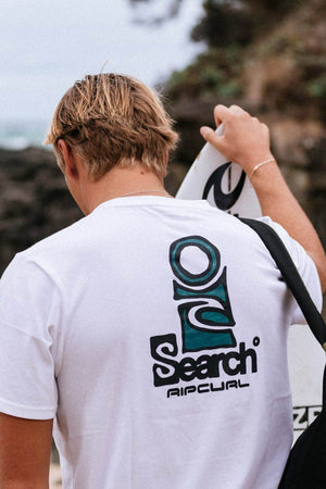 A person with blonde hair, wearing a white Rip Curl Vaporcool Search Stack Tee, is seen from behind holding a surfboard. The quick-drying t-shirt is perfect for an outdoor adventure near a rocky area with greenery.