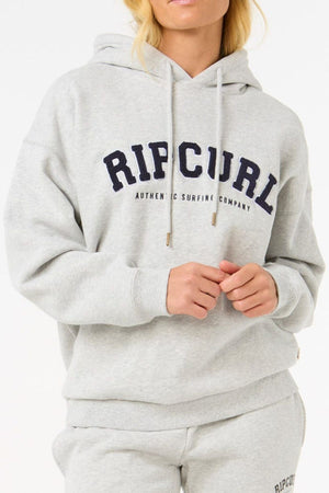 Wearing a light gray Rip Curl Varsity Hoodie made from soft Cotton Poly Fleece with an adjustable drawcord and bold chest logo, a person pairs it with matching sweatpants and stands against a plain background, showing only the lower half of their face.