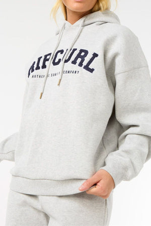 A person, face partially out of frame, wears a light gray Rip Curl Varsity Hoodie made from soft Cotton Poly Fleece with an adjustable drawcord and "RIP CURL AUTHENTIC SURF CO." in navy across the chest.