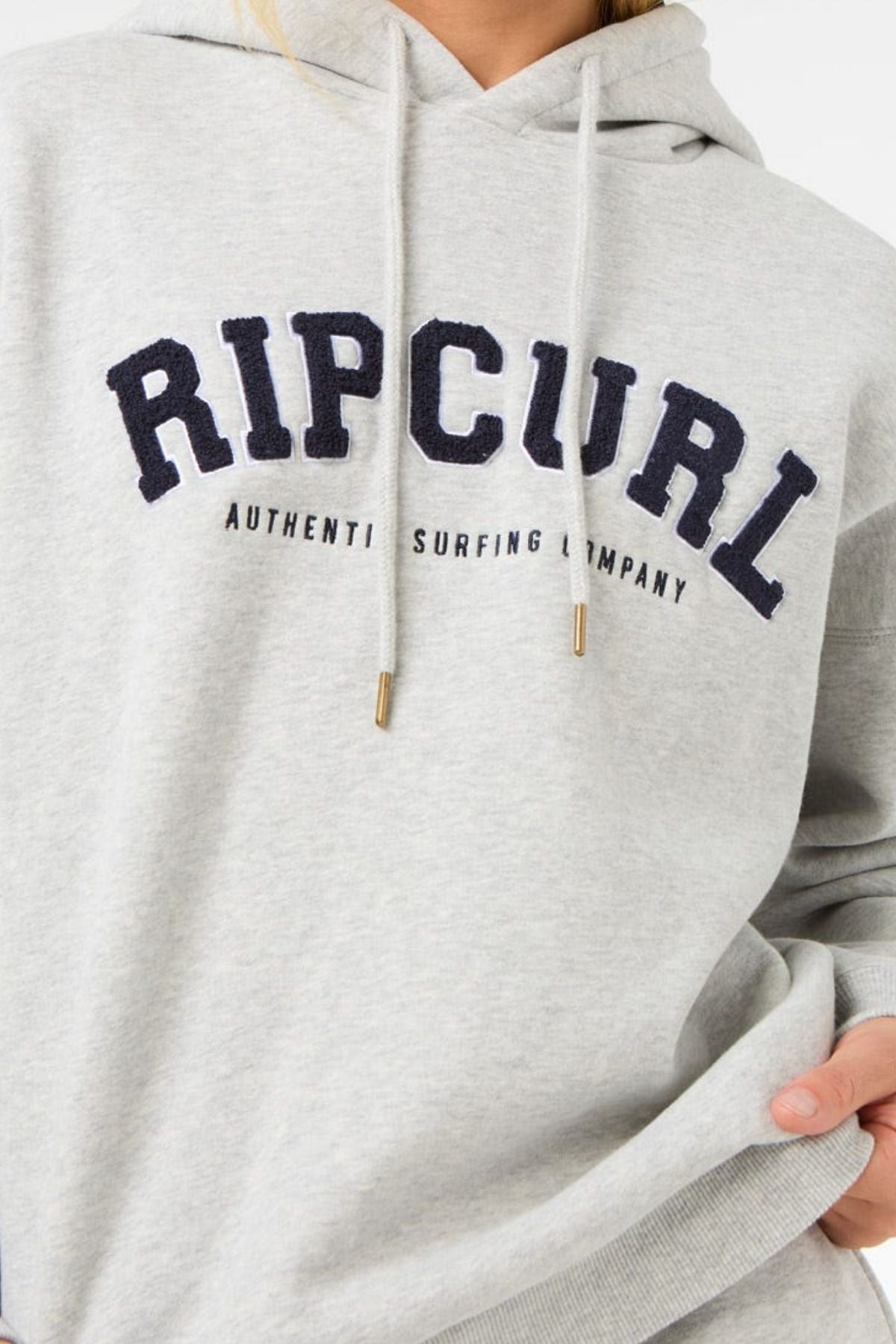 A person wears a light gray Rip Curl Varsity Hoodie made of soft Cotton Poly Fleece, with "RIP CURL AUTHENTIC SURFING COMPANY" on the front. Only their upper body and the hoodie’s adjustable drawcord hood are visible.