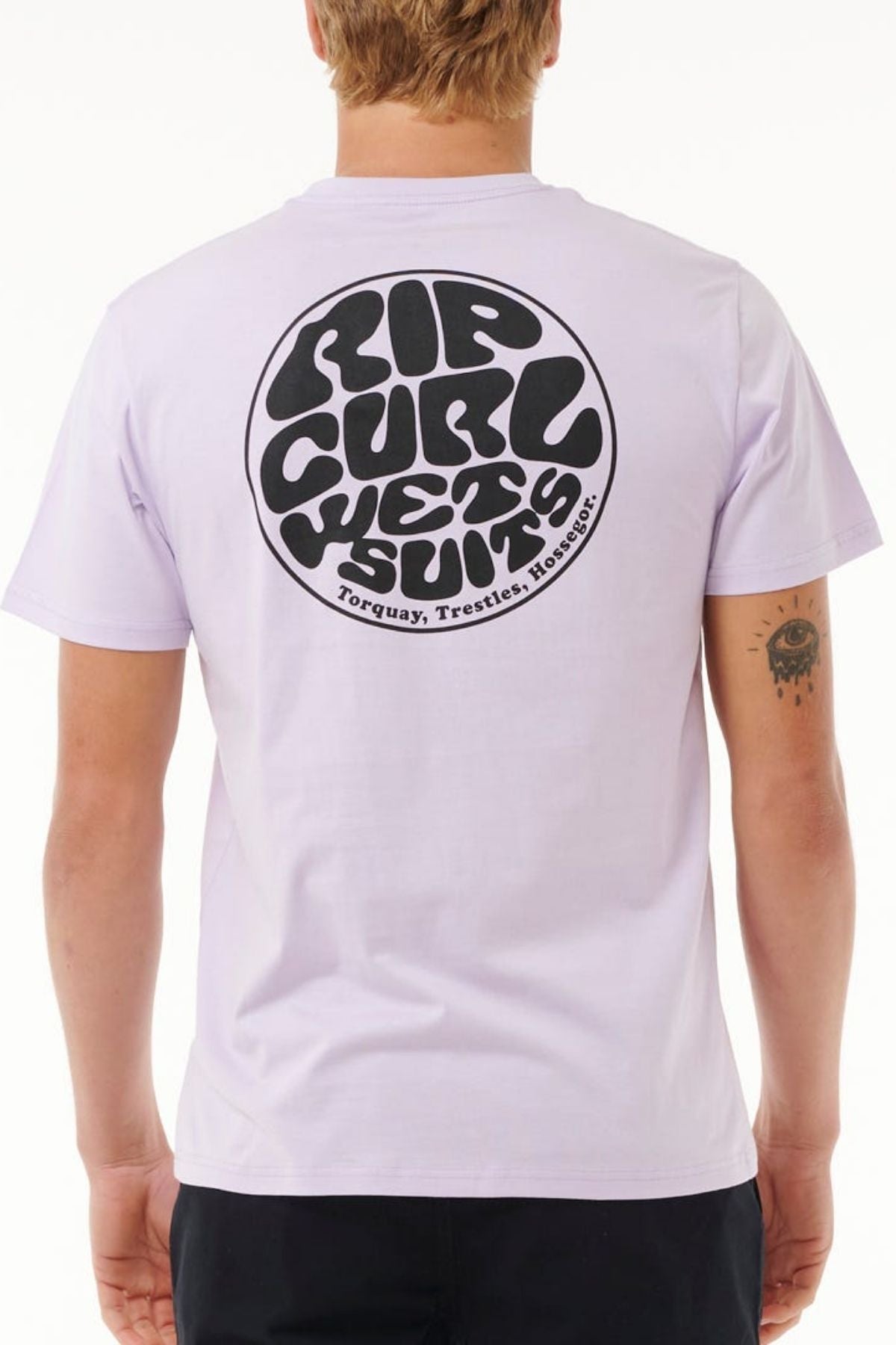 A person with short hair is wearing the Rip Curl Wetsuit Icon Tee, a light purple cotton shirt featuring a large circular logo on the back showcasing Torquay, Trestles, and Hossegor. They stand against a plain backdrop, embodying perfect style for outdoor activities.