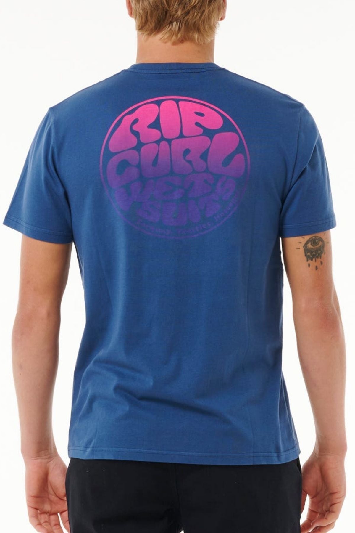 A blond-haired person is seen from behind in a blue Rip Curl Wetsuit Icon Tee with a round pink-purple logo on the back, made of lightweight cotton. They have a small tattoo on their left arm.