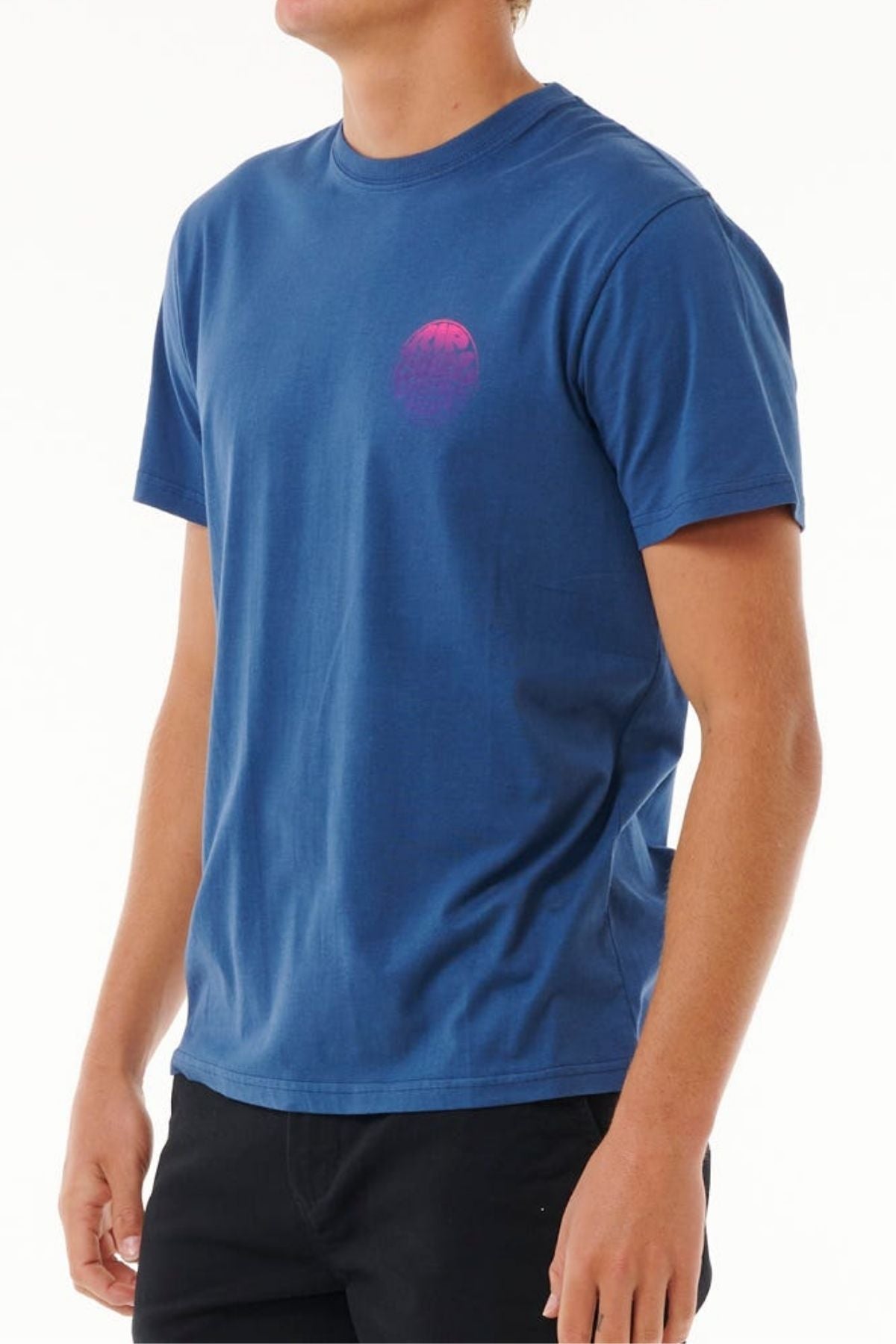 A person wearing the Rip Curl Wetsuit Icon Tee—a blue crew neck short-sleeve t-shirt with a small pink circular graphic on the left chest—paired with black pants, standing against a plain white background.