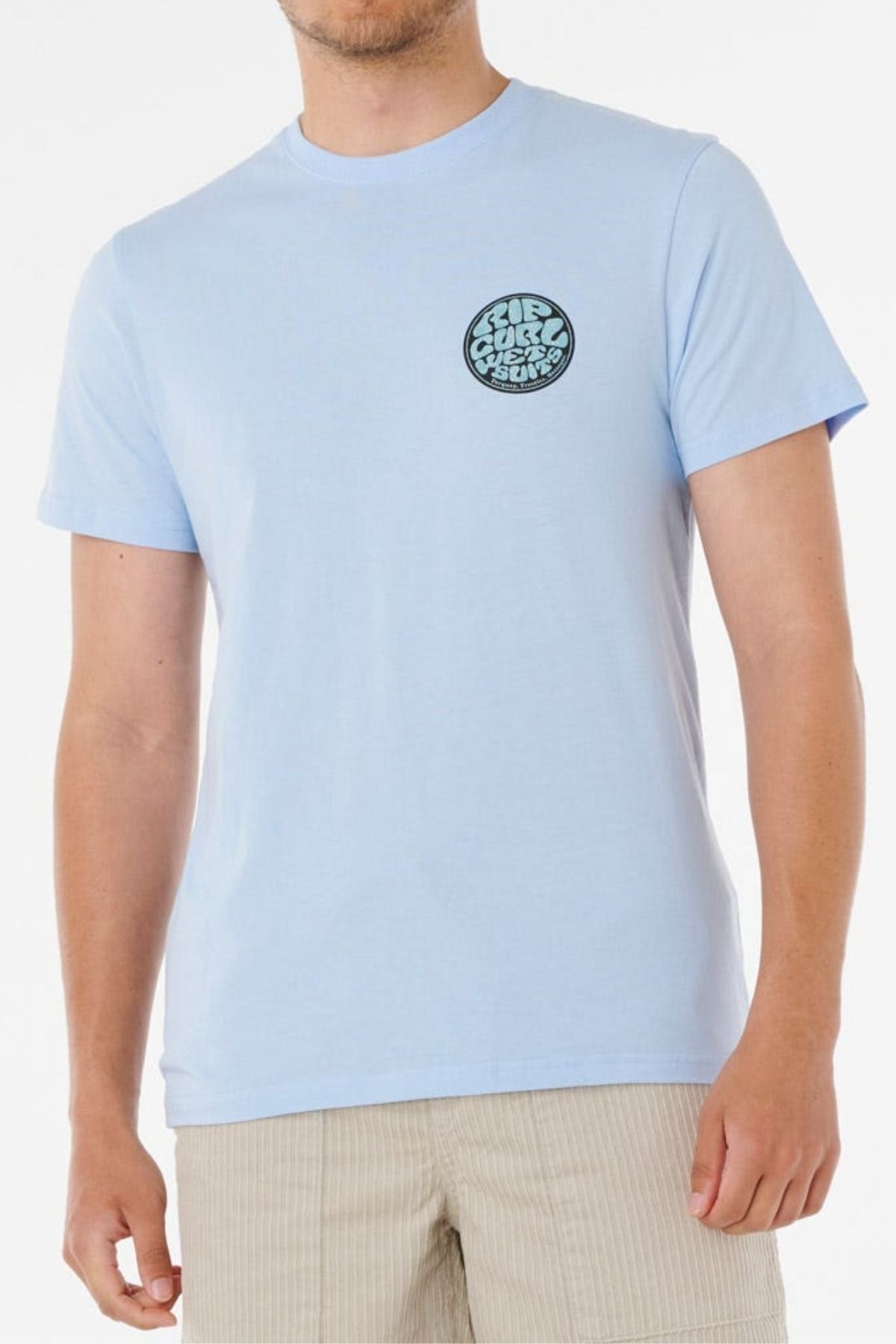A man wears the Rip Curl Wettie Passage Icon Tee, a light blue shirt with a round black and white "Surf Classic West Coast" logo on the left chest and beige shorts, creating an effortless, casual look against a plain white background.