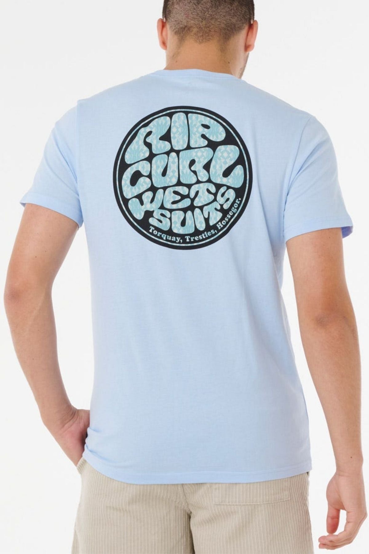 Someone wears the Rip Curl Wettie Passage Icon Tee in light blue, showcasing a large round black and white graphic on the back. The tee has a casual, comfortable fit as the person faces away from the camera.