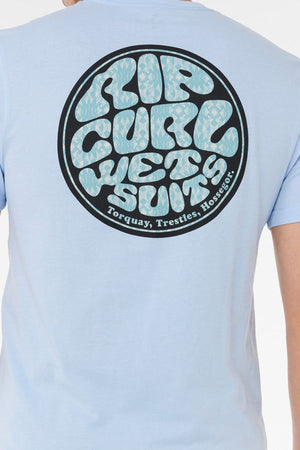 Wearing the Rip Curl Wettie Passage Icon Tee in light blue, featuring a large circular "RIP CURL WETSUITS" logo on the back with "Torquay, Trestles, Hossegor" below. This tee provides a comfortable fit for casual, laid-back style.