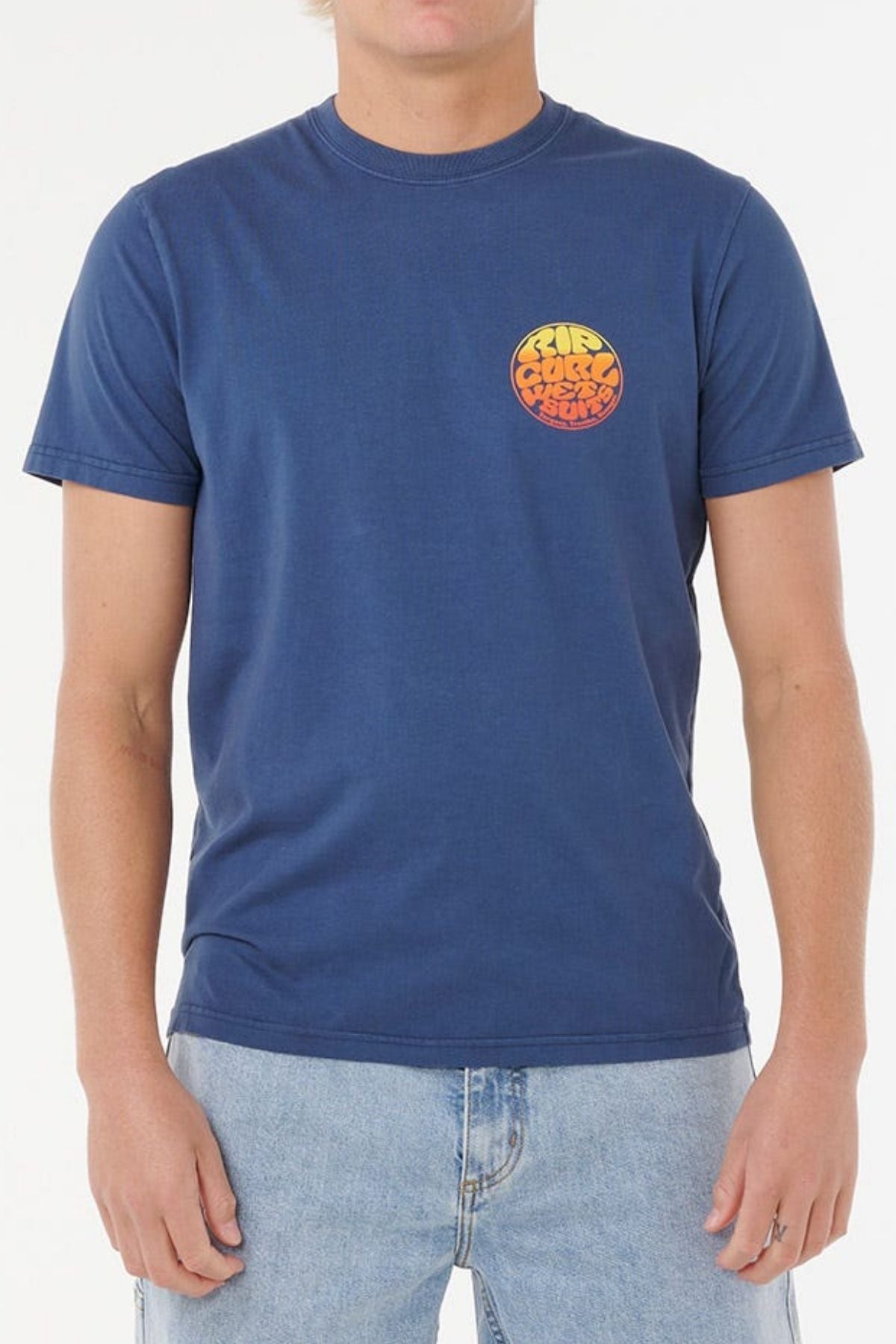 Wearing a Rip Curl Wettie Passage Icon Tee in blue with soft knit fabric and an orange-yellow chest graphic, paired with light blue jeans. The comfortable standard fit tee looks stylish, though the person’s face is partially out of frame.