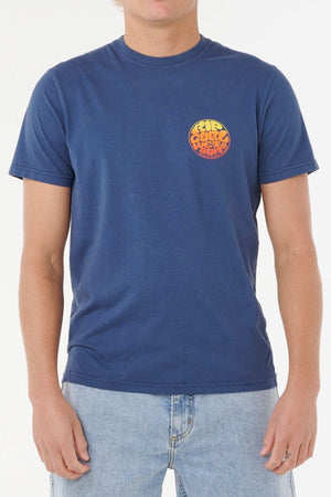 Wearing a Rip Curl Wettie Passage Icon Tee in blue with soft knit fabric and an orange-yellow chest graphic, paired with light blue jeans. The comfortable standard fit tee looks stylish, though the person’s face is partially out of frame.