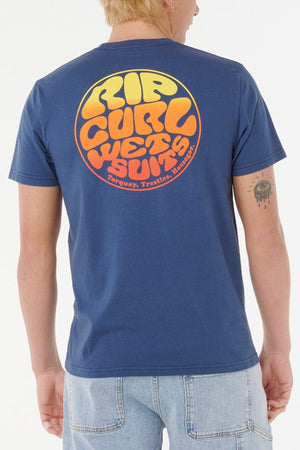 A person wearing a comfortable Rip Curl Wettie Passage Icon Tee in standard fit with soft knit fabric and a large retro-style "Rip Curl Wetsuits" back logo, paired with light blue jeans and sporting a small tattoo on their left arm.