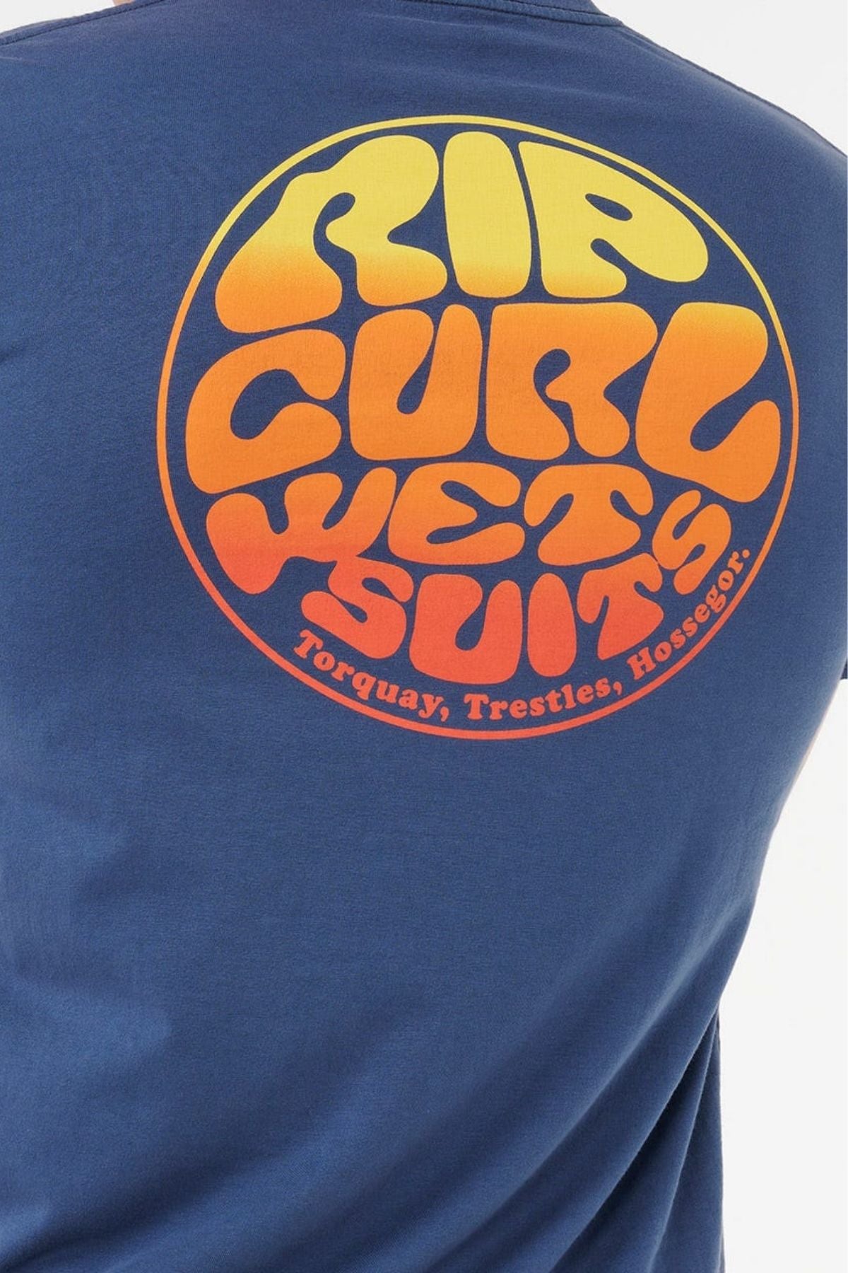 A person wearing a blue Rip Curl Wettie Passage Icon Tee with a round, retro "Rip Curl Wetsuits" logo in yellow and orange and "Torquay, Trestles, Hossegor" below. Crafted from soft knit fabric for a comfy standard fit.
