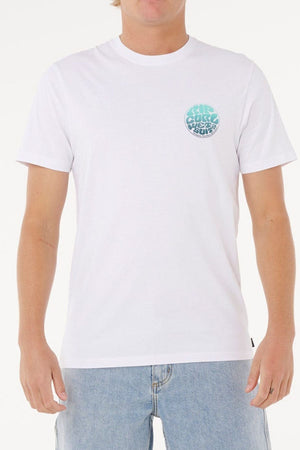 A person wears a Rip Curl Wettie Passage Icon Tee, a plain white short-sleeve shirt with a small blue and teal circular graphic on the left chest, made of soft knit fabric, paired with light blue jeans. The person's head is partially out of frame.
