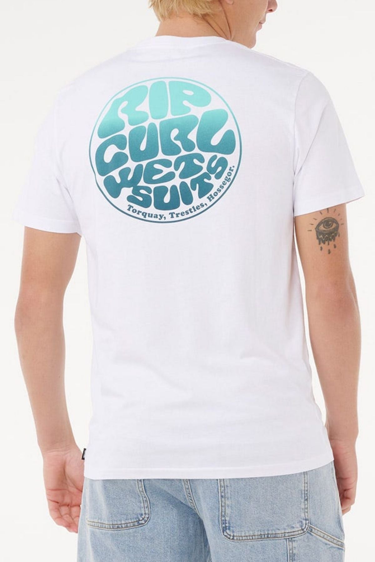 A person with short blond hair wears light blue jeans and a white Rip Curl Wettie Passage Icon Tee, made from soft knit fabric and featuring retro blue "Rip Curl Wetsuits" text on the back in a comfortable standard fit.