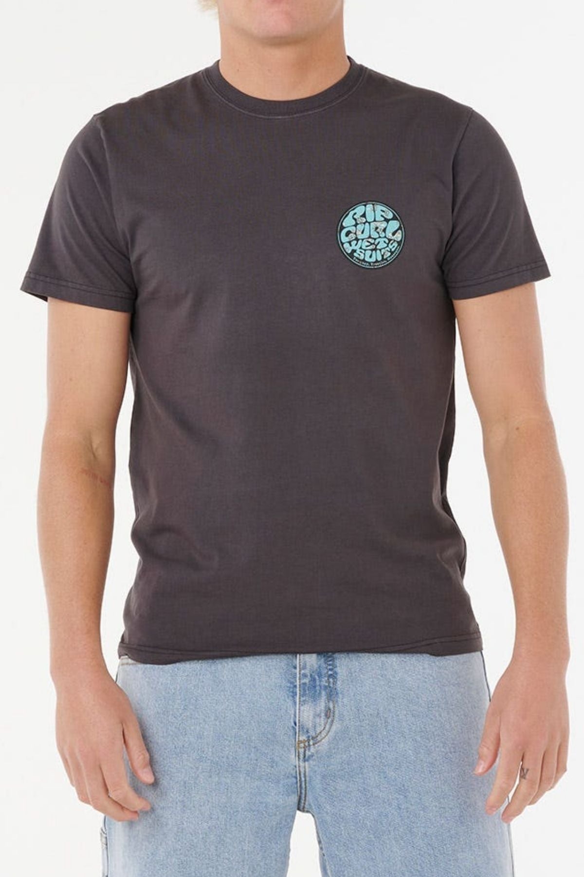 Wearing the Rip Curl Wettie Passage Icon Tee—a dark gray, short-sleeve shirt with a small blue and white circle graphic on the left chest—paired with light blue jeans for a casual look, against a plain white background.