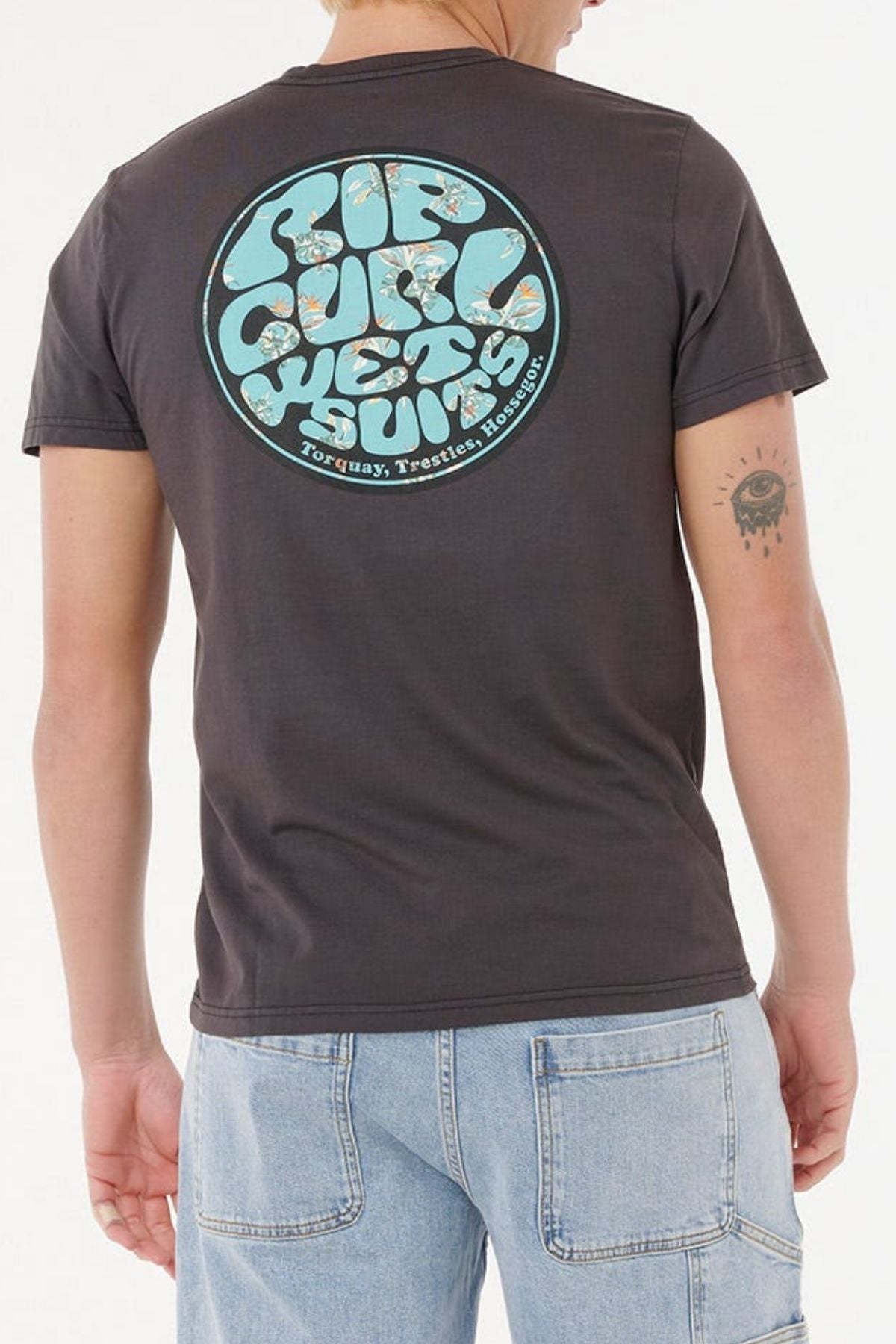 A person sports a casual look with the Rip Curl Wettie Passage Icon Tee—a dark shirt with a retro "Rip Curl Wet Suits" back graphic—paired with comfortable, standard-fit light blue denim jeans.