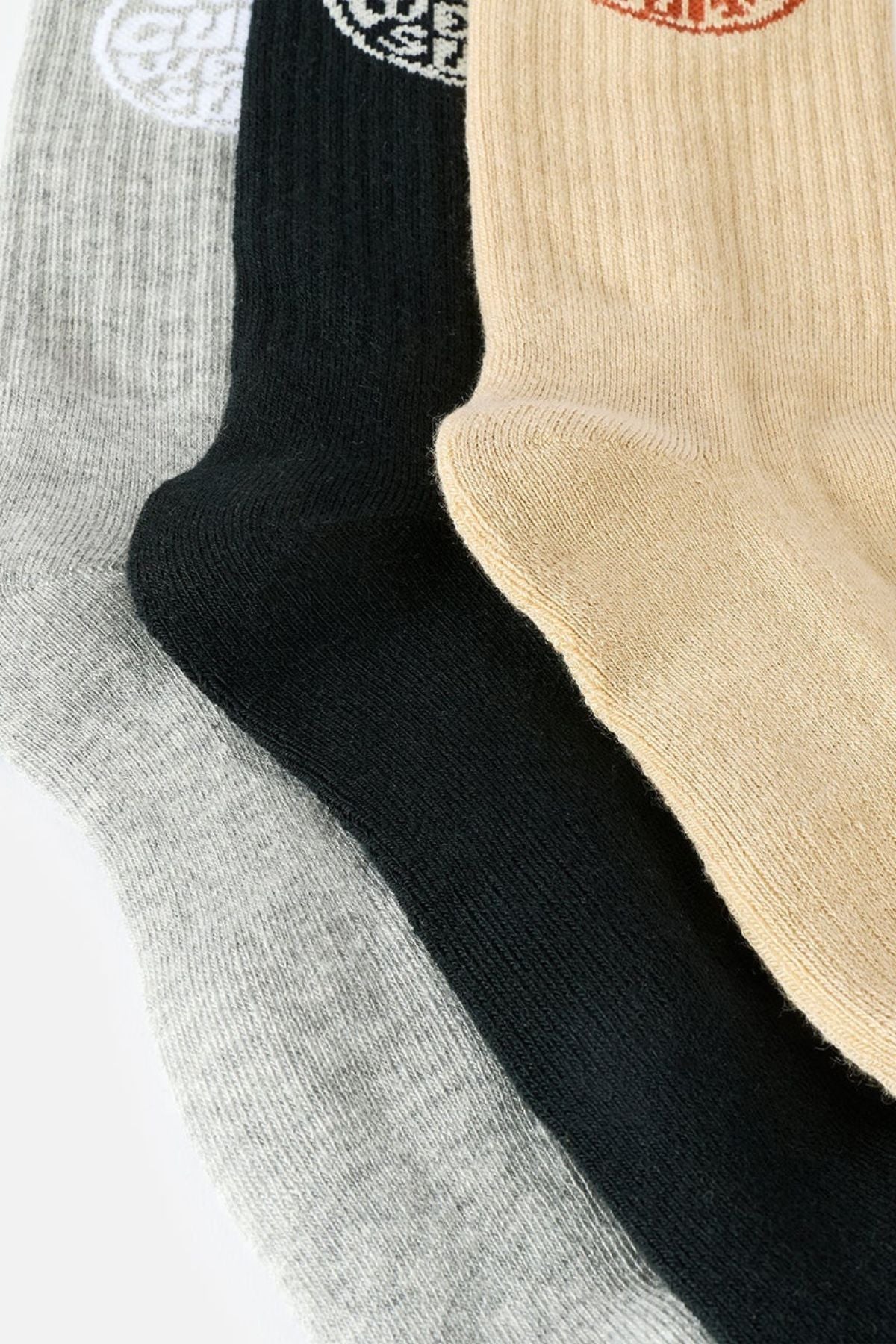 Three pairs of Rip Curl Wetty Crew Socks in light gray, black, and beige are laid out overlapping each other, each featuring a small circular emblem near the top. Designed with arch support and moisture-wicking properties, the Rip Curl Wetty Crew Sock 3 Pack offers both comfort and style for everyday wear.