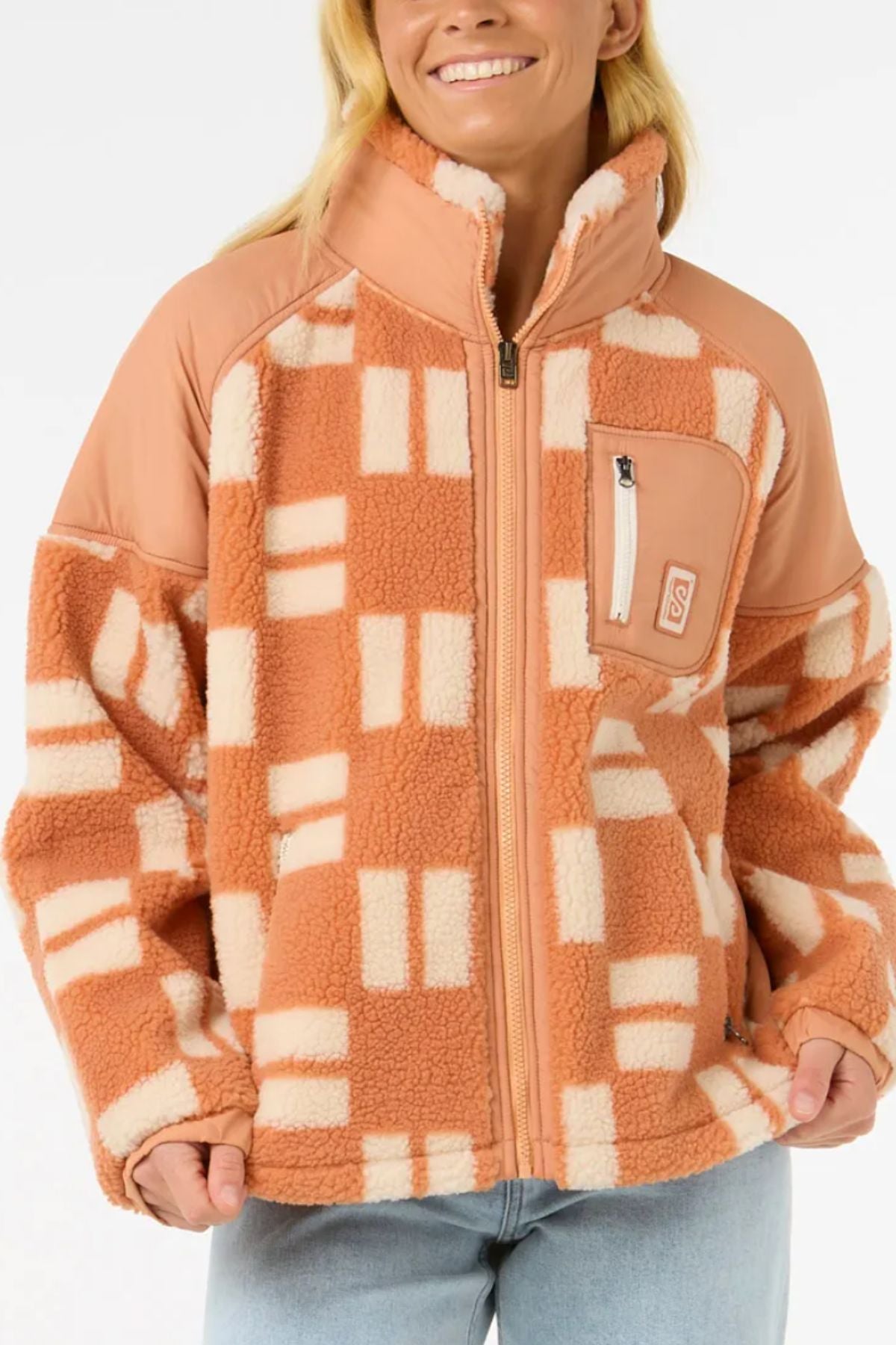 A person wearing the Rip Curl Anti-Series Shell Cove Fleece in a peach and cream checkered pattern with a high collar, zip front, and chest pocket pairs it with light blue jeans and smiles.