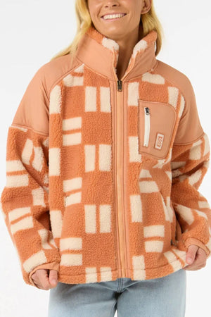 A person wearing the Rip Curl Anti-Series Shell Cove Fleece in a peach and cream checkered pattern with a high collar, zip front, and chest pocket pairs it with light blue jeans and smiles.