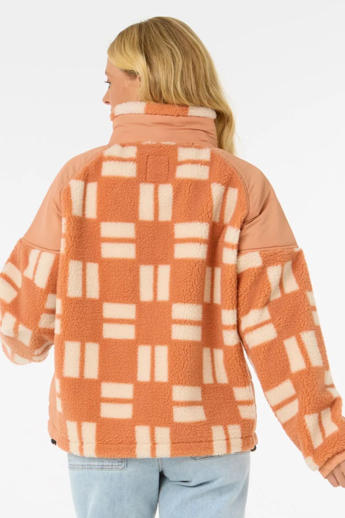 A person with long blonde hair, seen from behind, wears the Rip Curl Anti-Series Shell Cove Fleece featuring a peach and cream geometric pattern, solid peach panels on the shoulders and sleeves, and a relaxed fit, paired with light blue jeans.