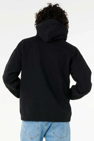 A person with curly hair, wearing a black Rip Curl Dosed Up Hood made from eco-friendly cotton polyester fleece and blue jeans, stands facing away from the camera against a plain light background.