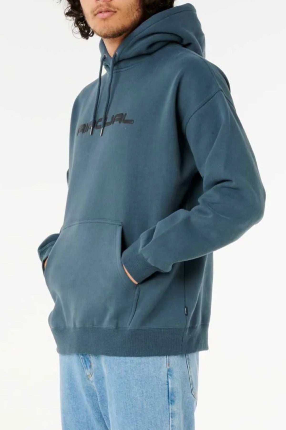 A person with curly hair wears a Rip Curl Dosed Up Hood made of cotton polyester fleece, featuring a front pocket and faded text, paired with light blue jeans, standing against a plain white background.
