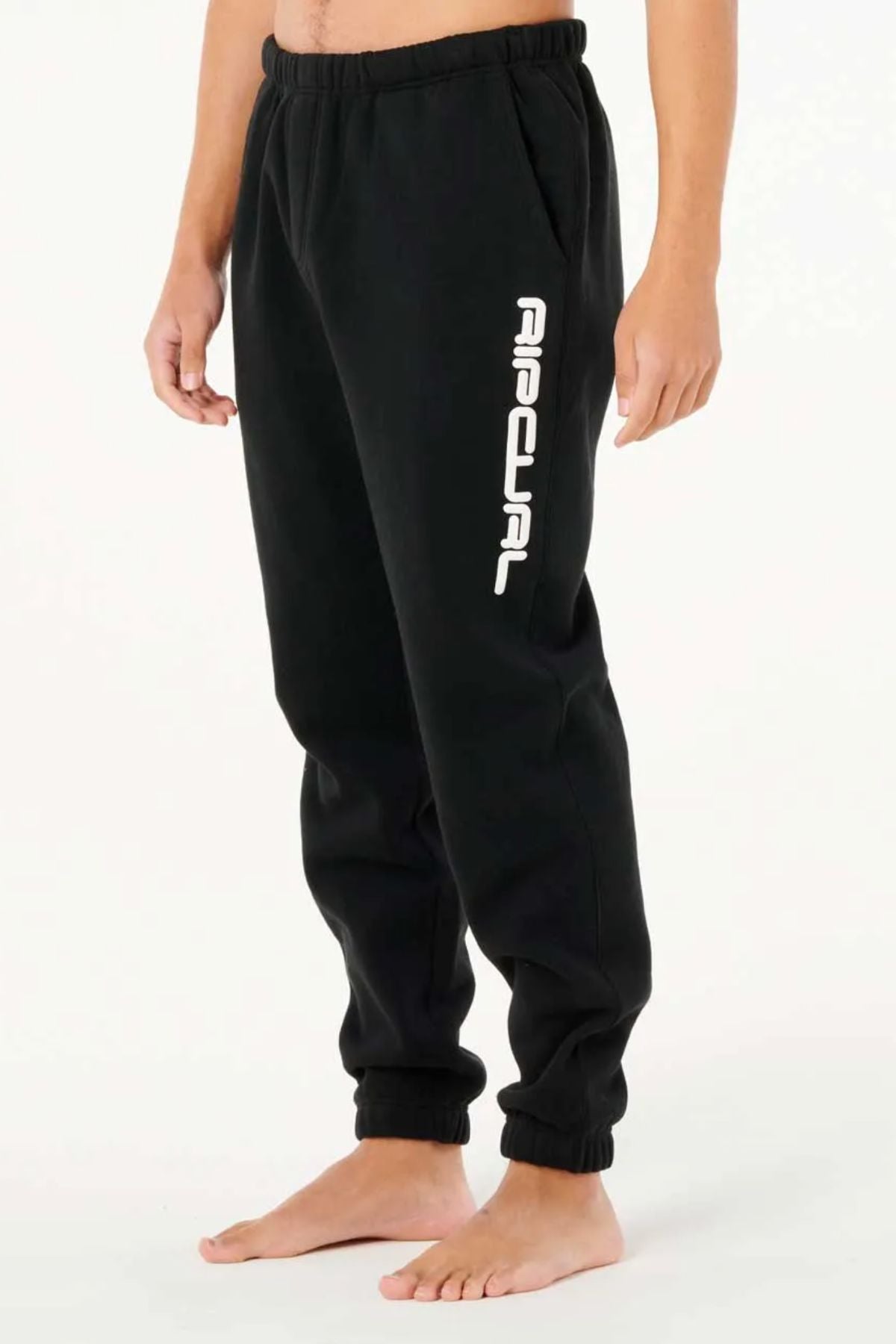 A person stands barefoot in black Rip Curl Dosed Up Trackpant joggers with elastic cuffs and bold white "ripcurl" lettering down the left leg, set against a plain white background.