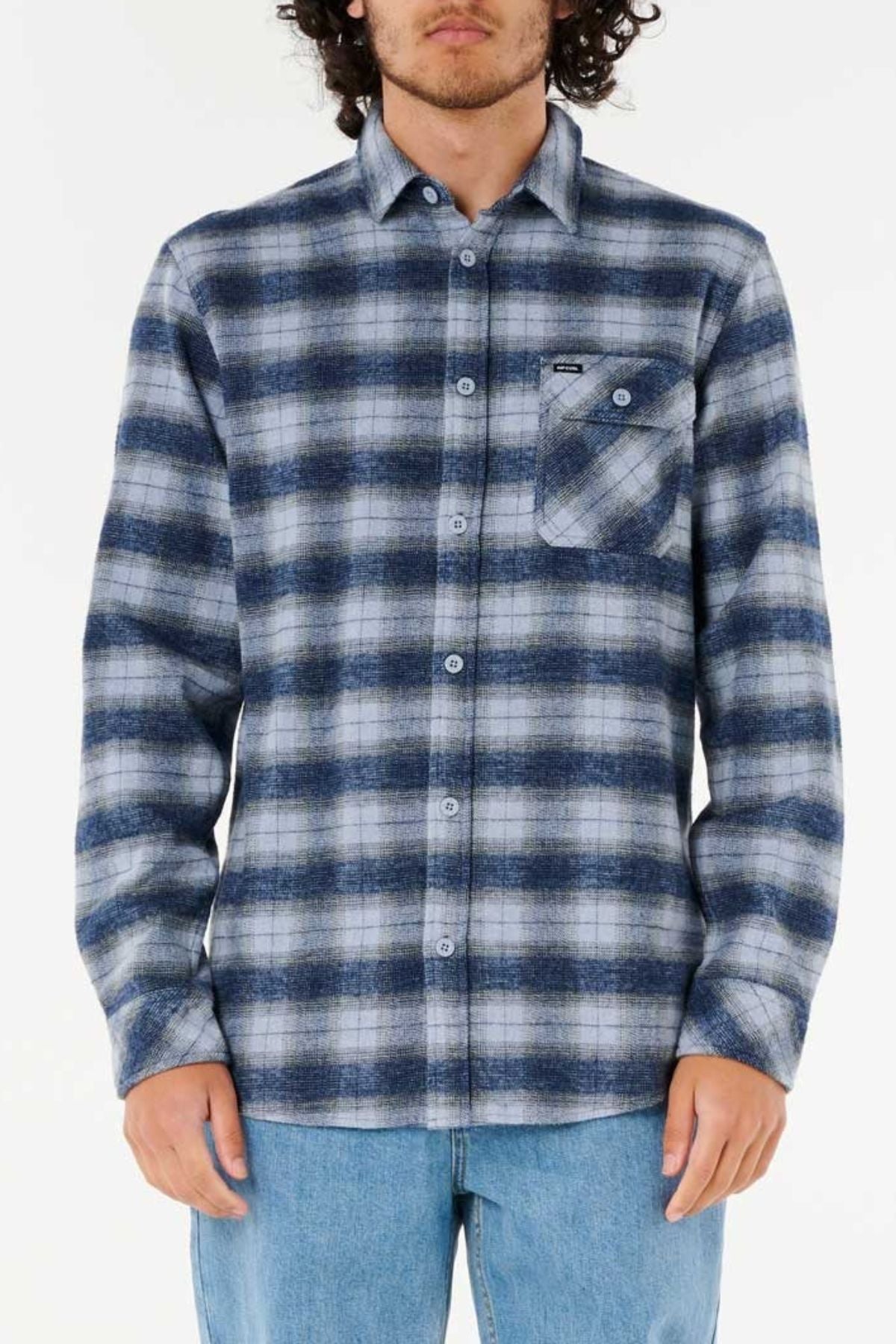 A person in a relaxed fit, blue and white plaid Rip Curl Grinners Flannel Shirt with a chest pocket stands before a plain background. Only the upper body is shown, with the face partially out of frame.