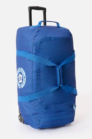 The Rip Curl Jupiter 80L Mixed Travel Bag is a large blue wheeled luggage with side straps, a retractable handle, and a white "Rising Rescue" flower logo, crafted from recycled polyester for durability and eco-friendly travel.