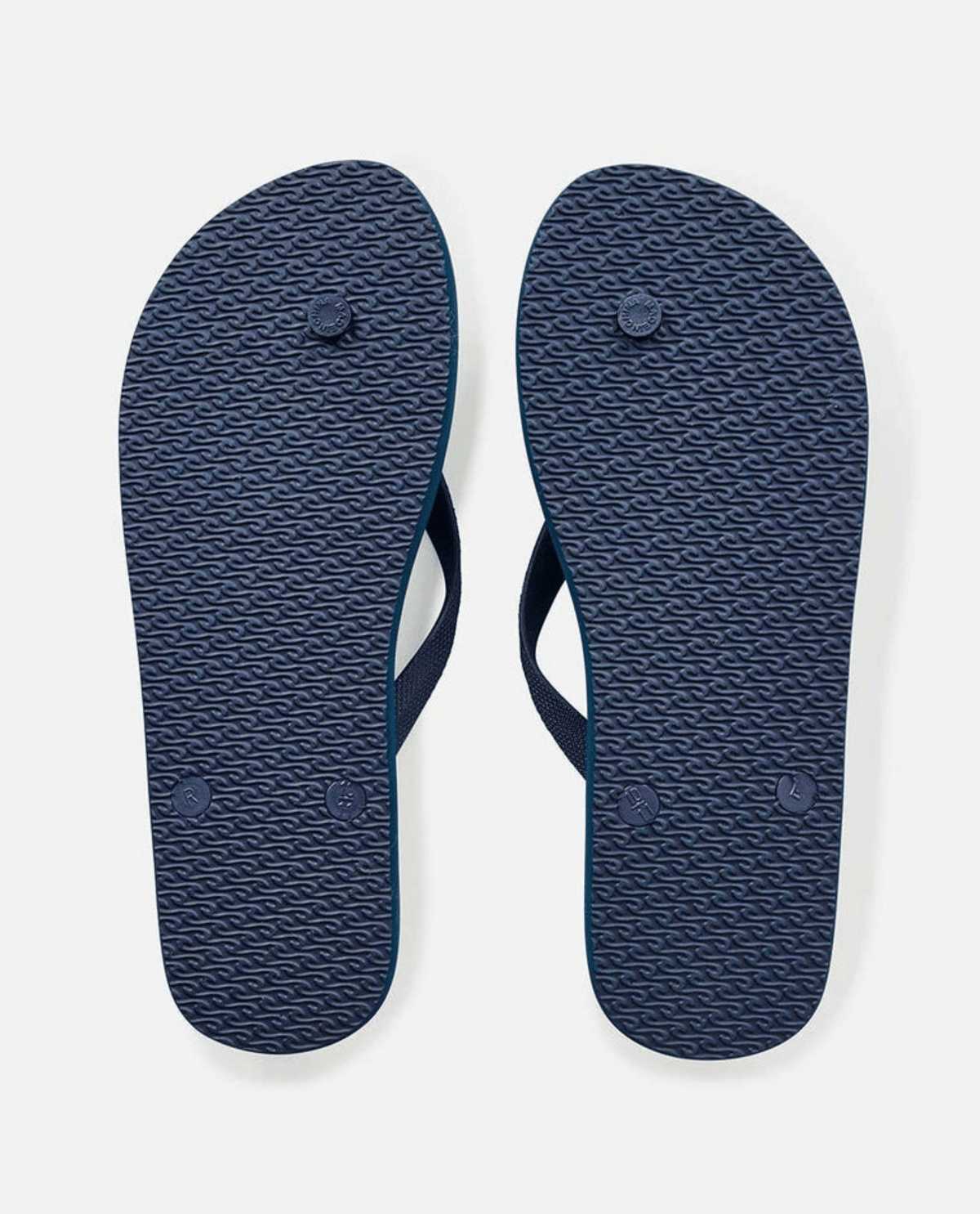 The image displays the bottoms of Rip Curl Brush Palm Bloom Thongs, featuring textured soles with circular grips and partially visible dark blue comfort straps. Made from BLOOM™ algae foam, these navy thongs offer eco-friendly style against a pristine white background.
