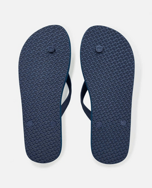 The image displays the bottoms of Rip Curl Brush Palm Bloom Thongs, featuring textured soles with circular grips and partially visible dark blue comfort straps. Made from BLOOM™ algae foam, these navy thongs offer eco-friendly style against a pristine white background.