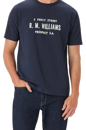 A person wears the RM Williams Bale T-Shirt in navy organic cotton, featuring a "5 Percy Street R. M. Williams Prospect S.A." print. Paired with dark blue jeans, their face is not shown.