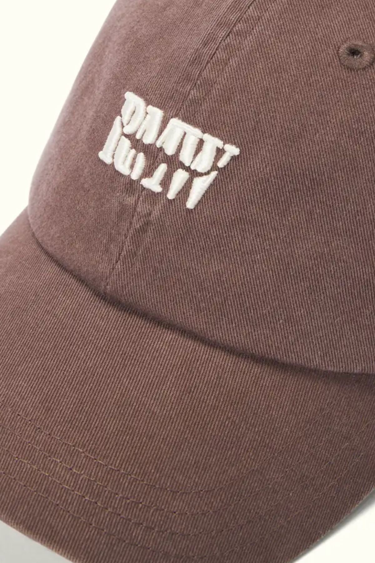 A close-up of the RM Williams Branded Twill Cap in brown organic cotton, featuring clear white embroidered text (upside down and distorted), visible stitching, textured fabric, and an adjustable strap.