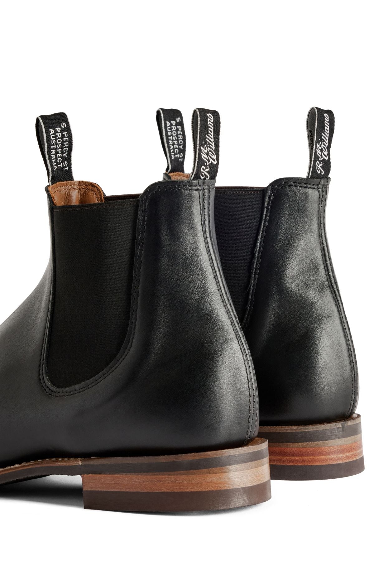 A close-up of two black RM Williams Comfort Craftsman Boots with brown soles shows "R.M. Williams" pull tabs and elastic side panels. The boots are placed heel to heel on a white background.