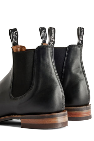 A close-up of two black RM Williams Comfort Craftsman Boots with brown soles shows "R.M. Williams" pull tabs and elastic side panels. The boots are placed heel to heel on a white background.