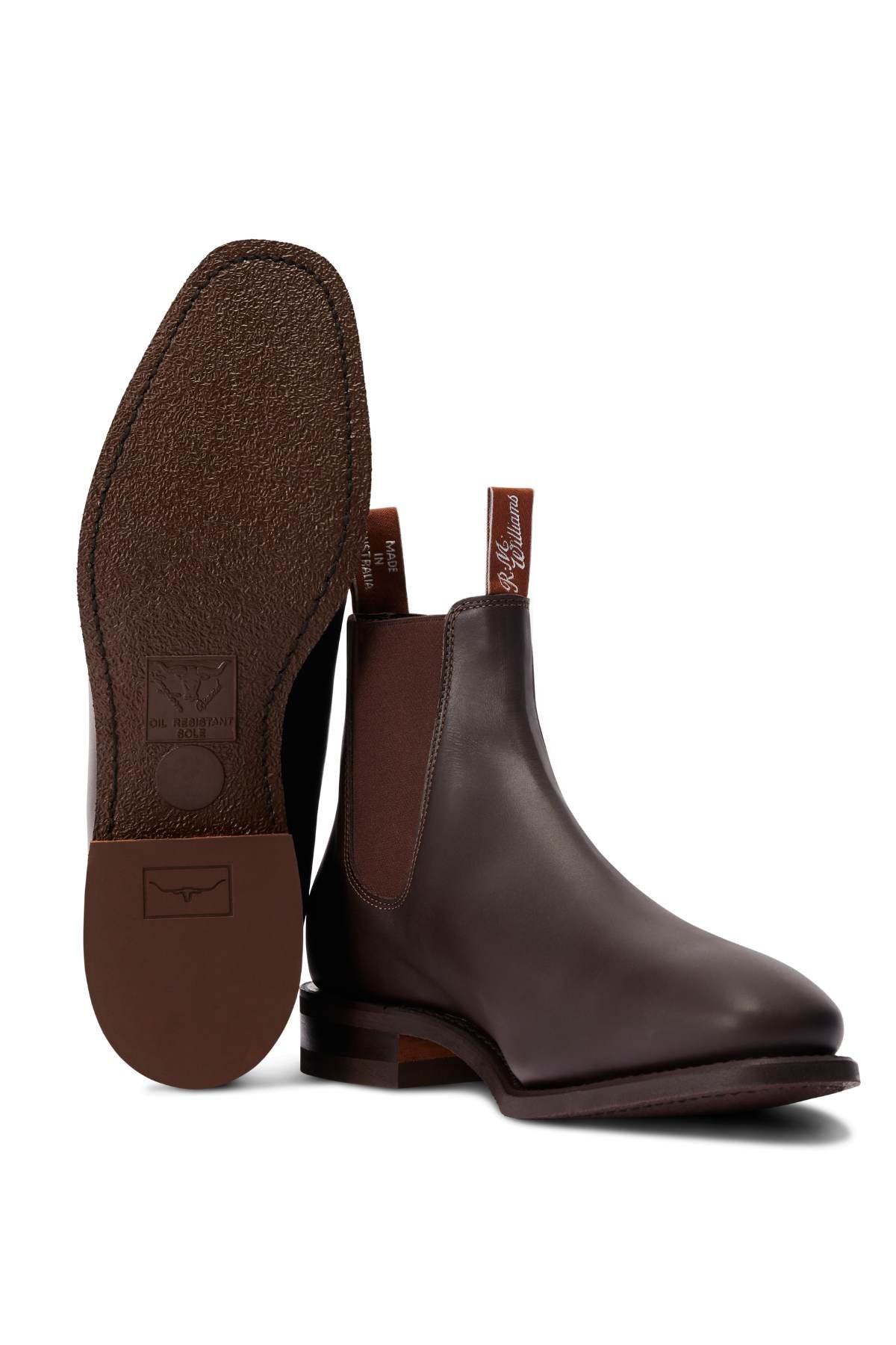 A pair of brown RM Williams Comfort Craftsman Boots featuring handcrafted leather, elastic side panels, and pull tabs; one boot stands upright while the other lies on its side to display the textured, branded sole.