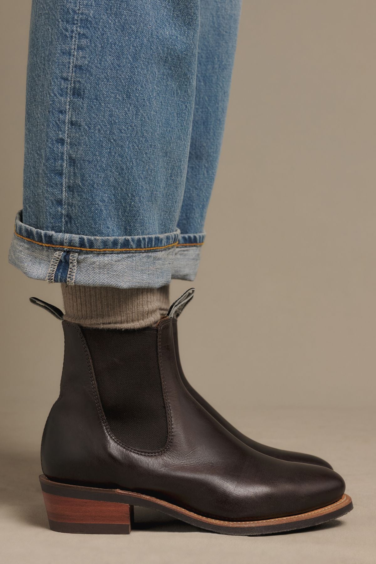 A person wearing blue cuffed jeans, beige socks, and RM Williams Comfort Lady Yearling Rubber Sole Boots in dark brown leather with a block heel stands on a neutral background.