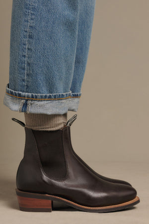 A person wearing blue cuffed jeans, beige socks, and RM Williams Comfort Lady Yearling Rubber Sole Boots in dark brown leather with a block heel stands on a neutral background.