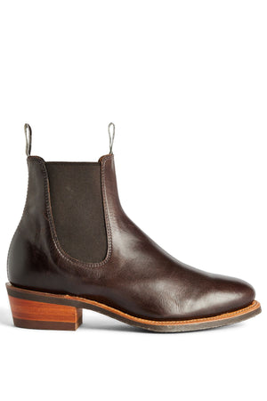 The RM Williams Comfort Lady Yearling Rubber Sole Boot is a dark brown leather Chelsea boot for women with black elastic side panels, a back pull tab, and a stacked wooden block heel, made in Australia and shown in side profile on white.