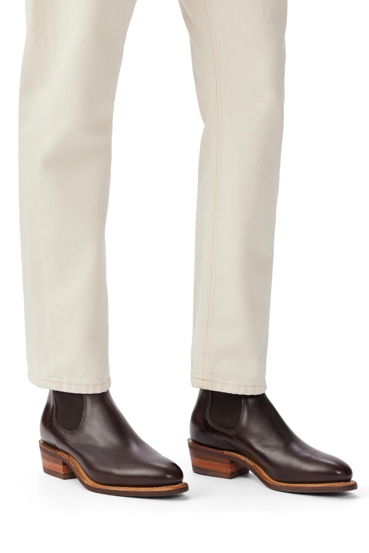 A person wearing off-white pants and RM Williams Comfort Lady Yearling Rubber Sole Boots in dark brown pull-up leather with wooden heels and Goodyear welt construction stands on a white background.