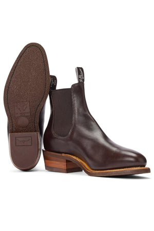 The RM Williams Comfort Lady Yearling Rubber Sole Boot is a pair of dark brown Chelsea boots in pull-up leather with elastic side panels, pull tabs, a textured rubber sole, wooden stacked heel, and Goodyear welt construction. One boot is upright; the other shows the sole.