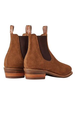 The RM Williams Comfort Suede Lady Yearling Rubber Sole Boot is shown from a rear side angle, featuring brown Italian suede, pull tabs, elastic side panels, stacked wooden heels, and rounded toes.