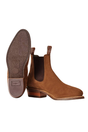 The RM Williams Comfort Suede Lady Yearling Rubber Sole Boot in brown Italian suede features elastic side panels and pull tabs. One boot stands upright, while the other shows off the textured sole and heel design.