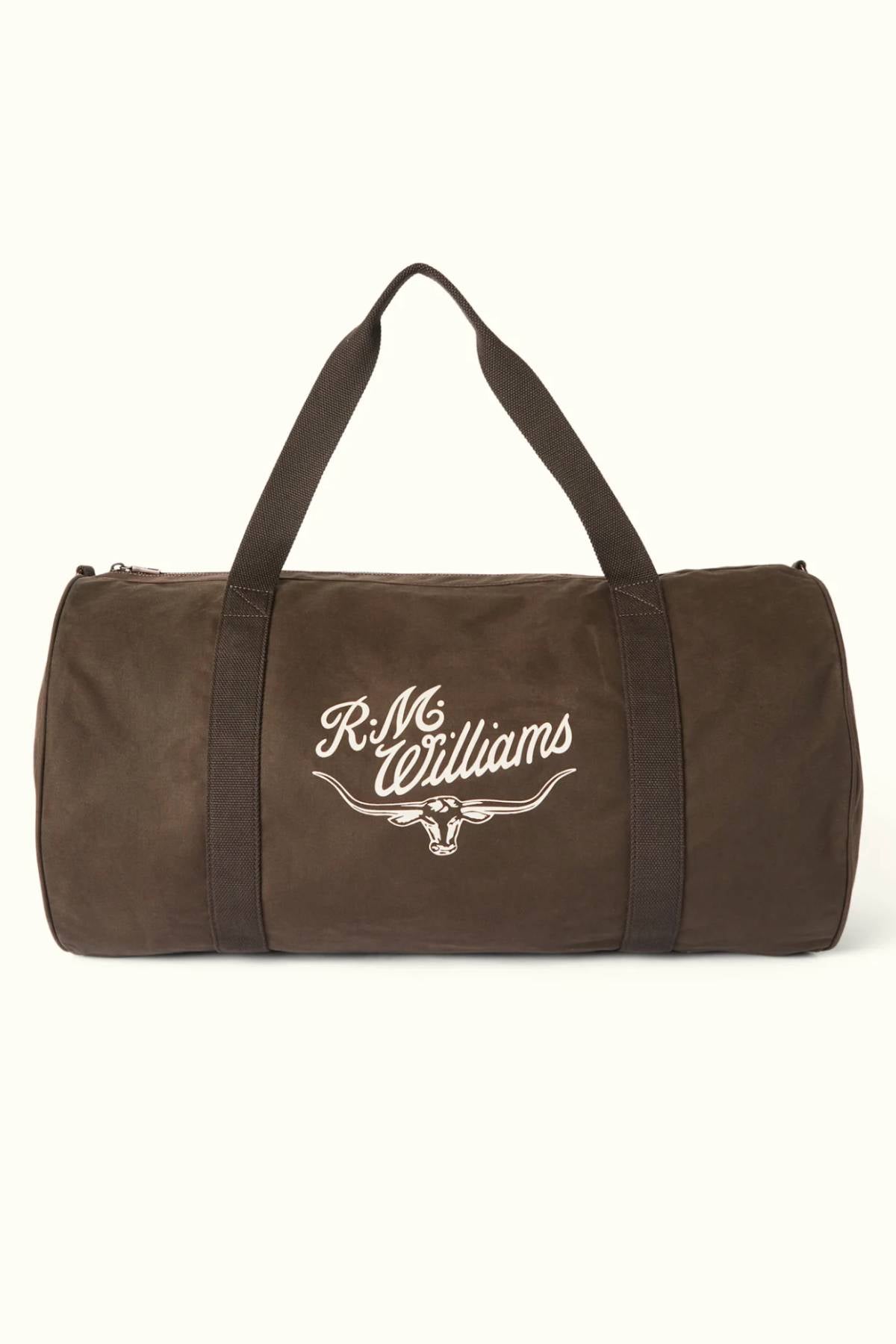 The RM Williams Ditty Bag is a brown cylindrical duffel made from waxed cotton canvas with black handles and "R.M. Williams" plus a bull logo printed in white on the side—an ideal travel companion.