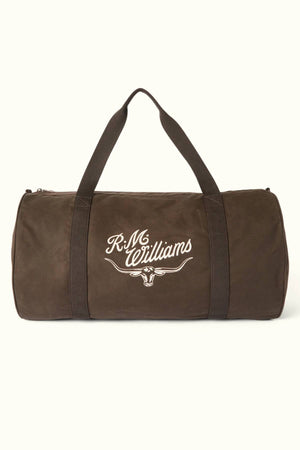 The RM Williams Ditty Bag is a brown cylindrical duffel made from waxed cotton canvas with black handles and "R.M. Williams" plus a bull logo printed in white on the side—an ideal travel companion.