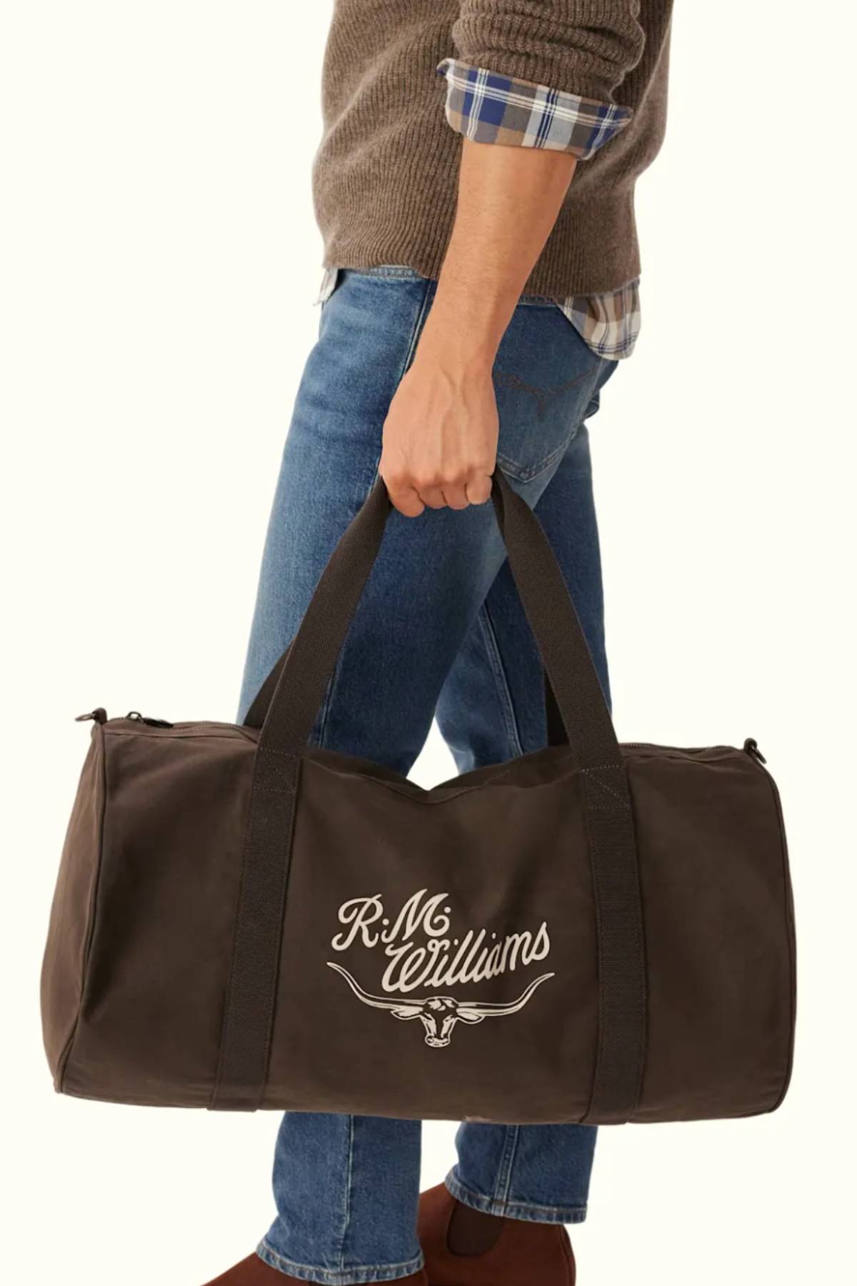 A person’s torso and legs are shown wearing a brown sweater, plaid shirt, and blue jeans while holding the RM Williams Ditty Bag—a black waxed cotton canvas duffel featuring "R.M. Williams" and a bull logo on its side.
