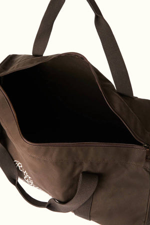 The RM Williams Ditty Bag is a brown duffel made from waxed cotton canvas with strong fabric handles and a small white graphic on the side. A partially open zipper reveals its spacious, empty interior—perfect as a travel companion.