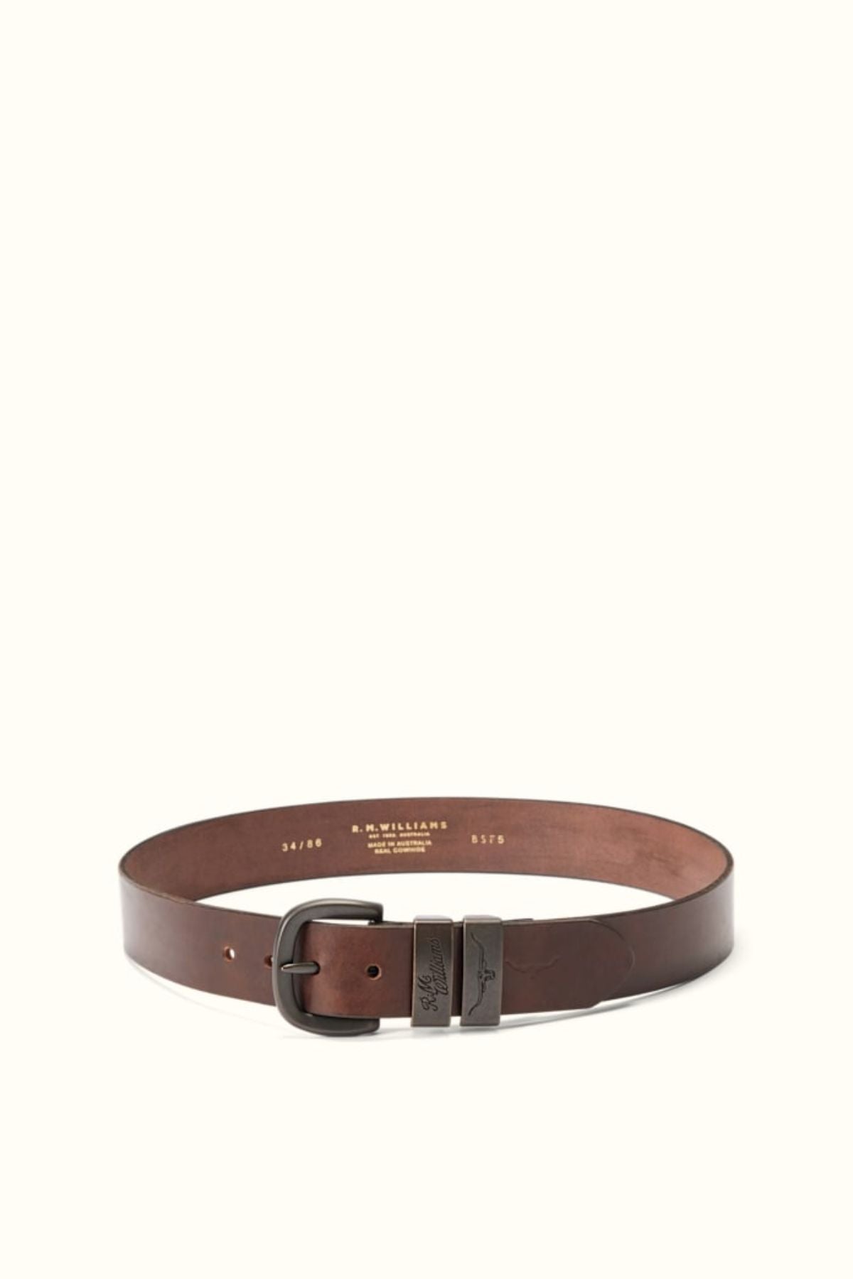 The RM Williams Drover Belt, a brown leather design with double keepers and a dark metal buckle, is shown in a circular shape on a light background. Gold embossed branding and size details appear on the inner side of the belt.