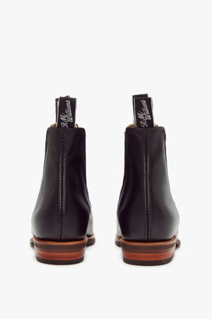 A pair of RM Williams Erica Boots in black full-grain leather, shown from the back with brown stacked heels and black pull tabs featuring white cursive text, displayed on a white background.