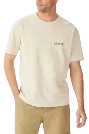 A man wears the RM Williams Flinders T-Shirt—a cream, relaxed-fit organic cotton jersey with small left chest text—paired with khaki shorts. He stands with one hand in his pocket, gazing slightly to the side.